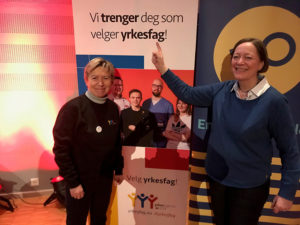 F.v.: Elisabeth Lange, sekretariatsleder i WorldSkills Norway og Bente Søgaard, seniorrådgiver i YS, peker på et skilt der det står «Vi trenger deg som velger yrkesfag»
